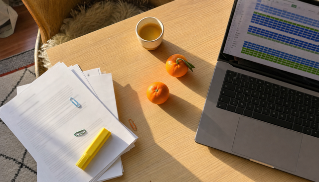 Vue plongeante : pile de documents papier et ordinateur affichant un tableur coloré, trombones et clémentines au soleil