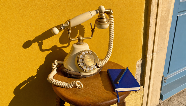 Téléphone à cadran vintage crème, combiné soulevé, sur fond jaune mimosa au soleil, carnet bleu roi à côté